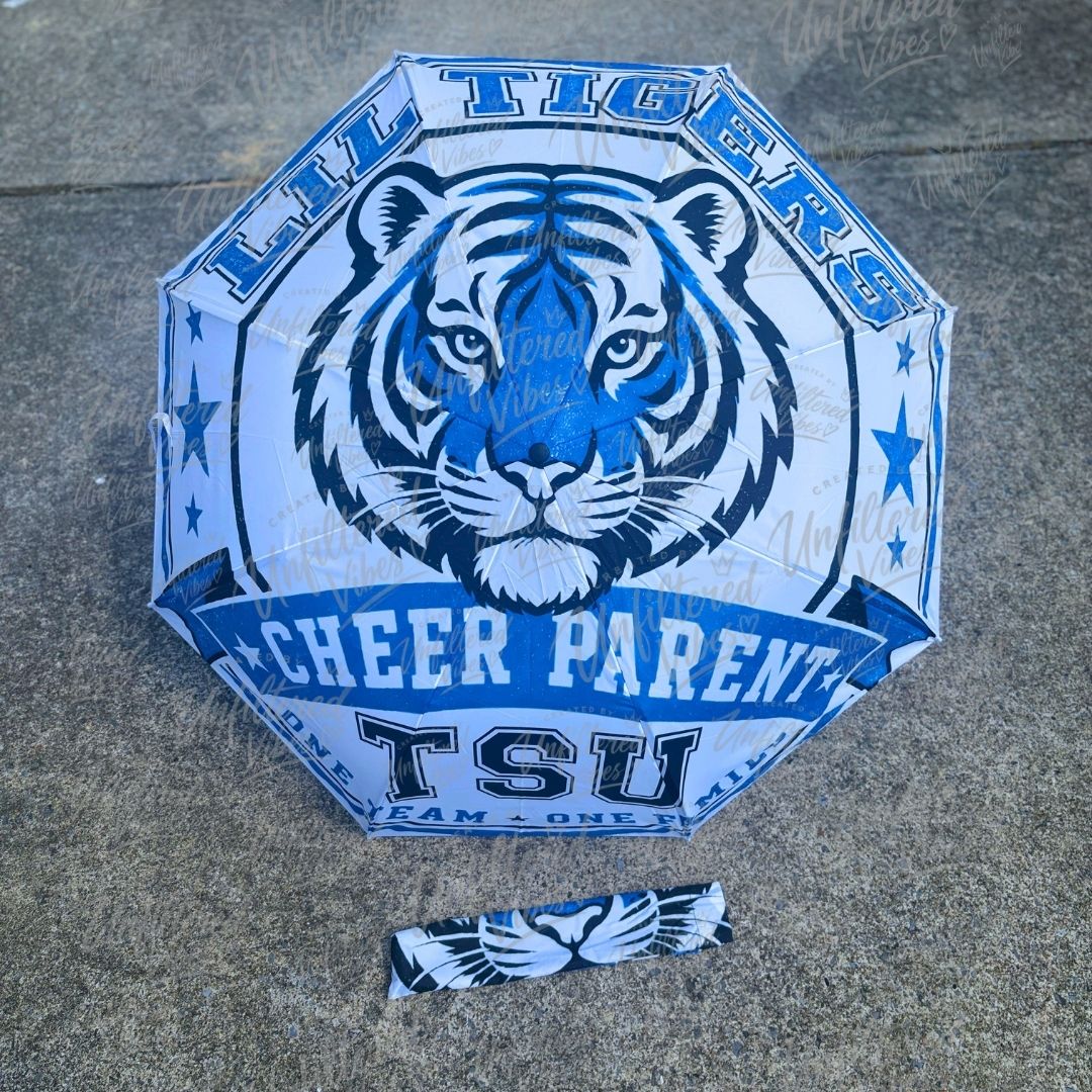 A blue and white octagonal umbrella laid flat on a gray concrete surface, featuring a large tiger head graphic and the text 'Lil Tigers Cheer Parent TSU.' A matching small accessory piece with a white tiger face and blue wings is placed below the umbrella.